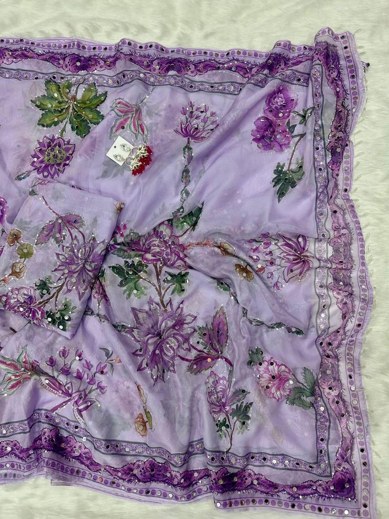 May include: A light purple floral scarf with a matching pouch. The scarf features a repeating pattern of purple flowers and green leaves. The edges are trimmed with a dark purple border and small silver embellishments. The fabric appears to be lightweight and flowing.
