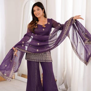 Puede incluir: Conjunto de dos piezas color ciruela, compuesto por pantalón de pernera ancha y top túnica. La túnica y el pantalón están adornados con bordados dorados. El conjunto incluye una bufanda transparente a juego con ribetes dorados. Pendientes dorados.