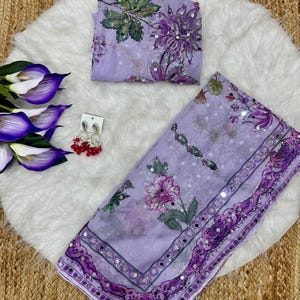 May include: A light purple saree with floral patterns and mirror accents. The saree is accompanied by a matching blouse piece. A pair of red and silver earrings and artificial flowers are also present.
