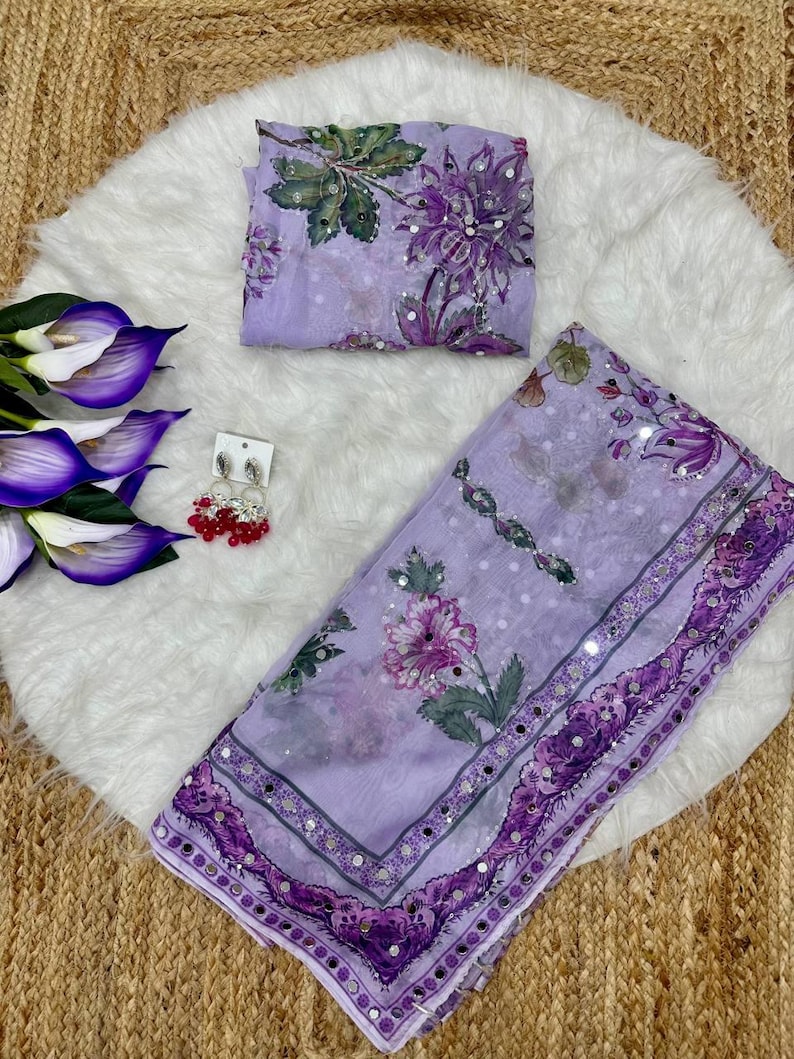 May include: A light purple saree with floral patterns and mirror accents. The saree is accompanied by a matching blouse piece. A pair of red and silver earrings and artificial flowers are also present.