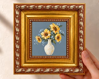 Pode incluir: Uma pintura quadrada numa moldura decorativa dourada e vermelho-escura com uma representação a óleo de girassóis amarelos e pequenas flores azuis num vaso branco sobre um fundo azul ardósia com textura de pinceladas.