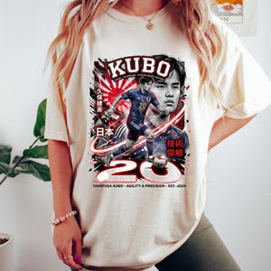 May include: Cream-colored t-shirt featuring a graphic of a soccer player with the name "KUBO" above. The design includes the number "20" and Japanese text. The text "TAK&Eacute;FUSA KUBO - AGILITY & PRECISION - EST. 2024" is also visible.