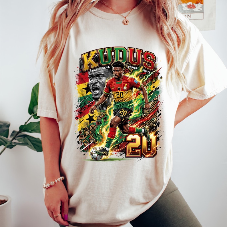 May include: A cream-colored t-shirt featuring a graphic of a soccer player in a red, yellow, and green uniform, with the text "KUDUS" and "20". The design incorporates the Ghana flag and other graphic elements.