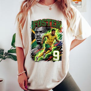 May include: Cream-colored t-shirt featuring a graphic of a soccer player in a yellow and green uniform, with the name "FOSTER" and "SOUTH AFRICA" in green. The number "9" is prominently displayed.