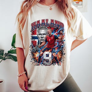 May include: Cream-colored t-shirt featuring a graphic of a soccer player with the name "HAALAND" in bold letters. The design includes the Norwegian flag, lightning bolts, and the number 9. The shirt has short sleeves.