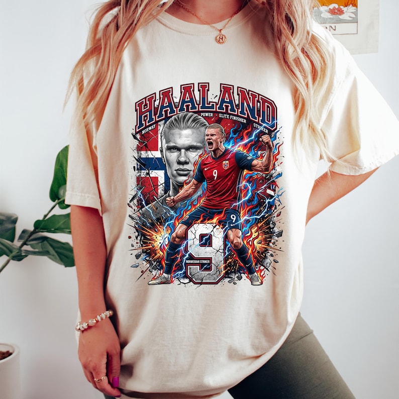 May include: Cream-colored t-shirt featuring a graphic of a soccer player with the name "HAALAND" in bold letters. The design includes the Norwegian flag, lightning bolts, and the number 9. The shirt has short sleeves.