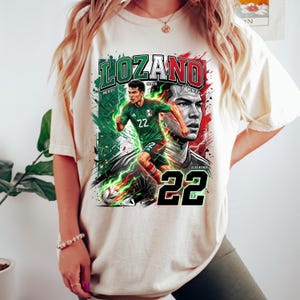 May include: Cream-colored t-shirt featuring a graphic of a soccer player in a green and red uniform, with the name "LOZANO" and the number "22". The design includes the Mexican flag colors and the words "MEXICO" and "WINNER".