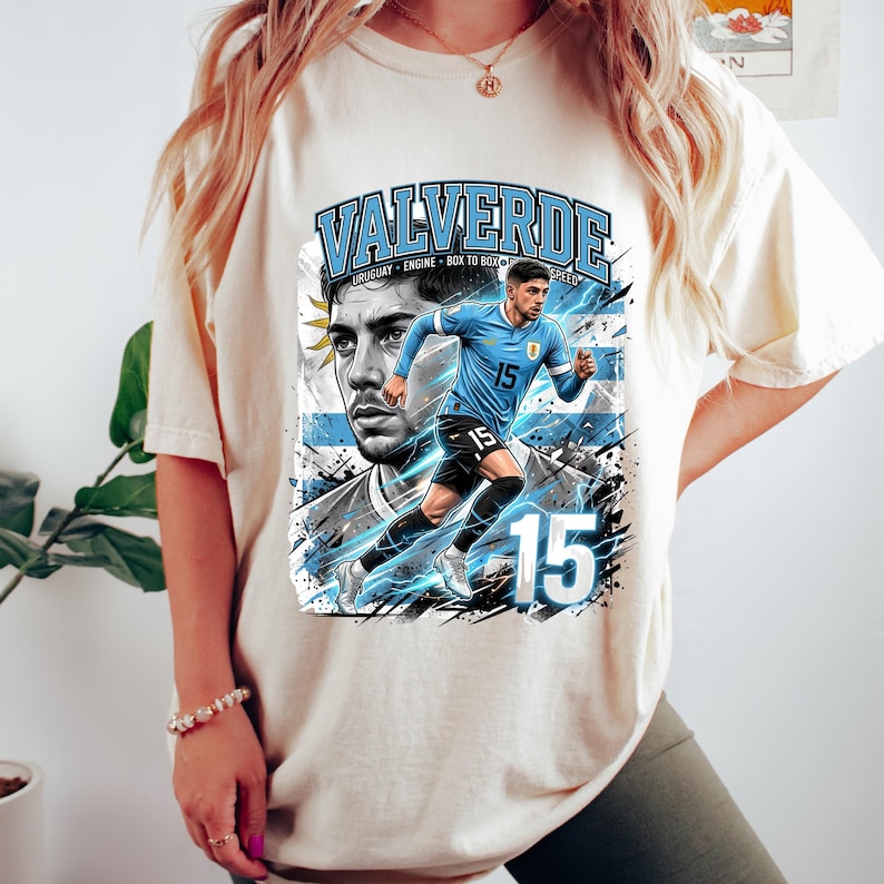 May include: Cream-colored t-shirt featuring a graphic of a soccer player in a blue uniform with the number 15. The design includes the name "VALVERDE" and the Uruguayan flag. The shirt has a relaxed fit.