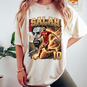 May include: Cream-colored t-shirt featuring a graphic of a soccer player in a red uniform with the name "SALAH" in gold lettering. The design includes the number 10 and the text "EGYPT KING OF EGYPT SPEED ELITE WINGER CAIRO."