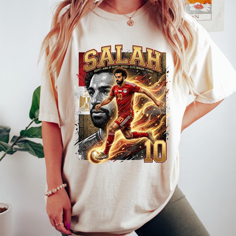 May include: Cream-colored t-shirt featuring a graphic of a soccer player in a red uniform with the name "SALAH" in gold lettering. The design includes the number 10 and the text "EGYPT KING OF EGYPT SPEED ELITE WINGER CAIRO."