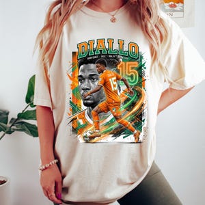 May include: Cream-colored t-shirt featuring a graphic of a soccer player in an orange uniform with the name "DIALLO" in green and orange. The design includes the number 15 and the words "IVORY COAST SKILL AGILITY STAR".