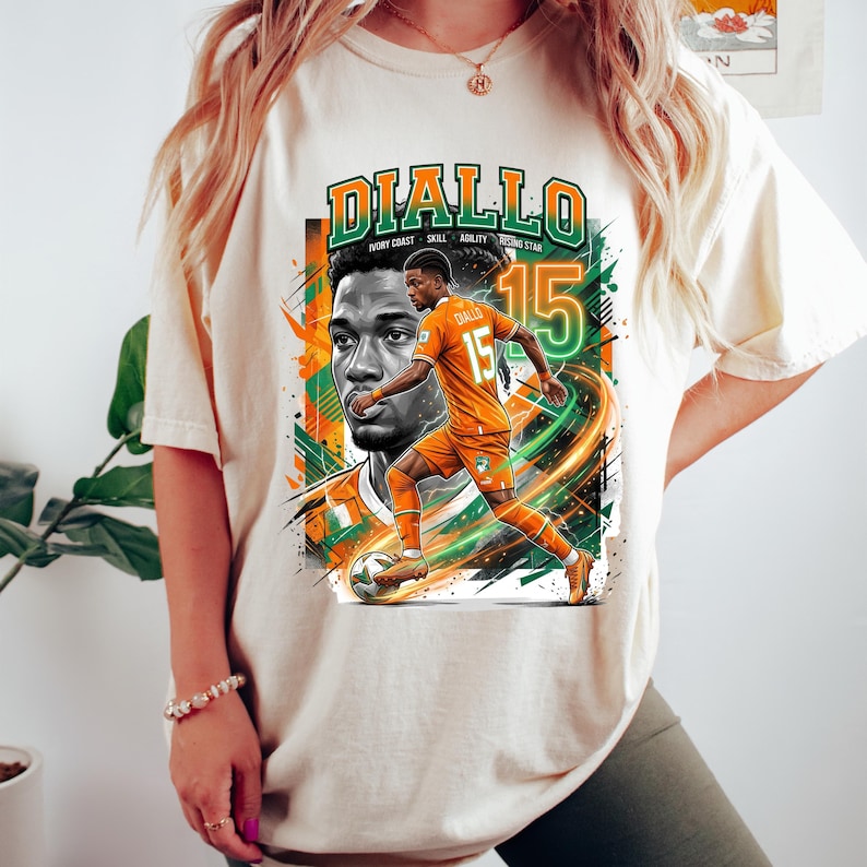 May include: Cream-colored t-shirt featuring a graphic of a soccer player in an orange uniform with the name "DIALLO" in green and orange. The design includes the number 15 and the words "IVORY COAST SKILL AGILITY STAR".