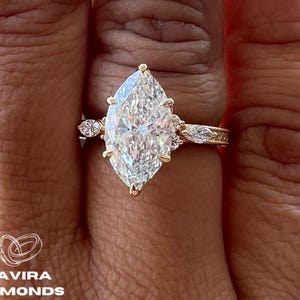 May include: A marquise-cut diamond engagement ring with a gold band. The ring features a large, clear center stone and smaller accent stones. The ring is on a hand with dark nail polish, with the text "D'AVIRA DIAMONDS".