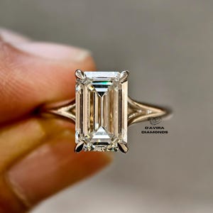 May include: An emerald-cut diamond ring featuring a large, rectangular center stone held by four prongs on a polished metal band with a split shank design. The text D'AVIRA DIAMONDS is printed in black near the base of the ring setting.