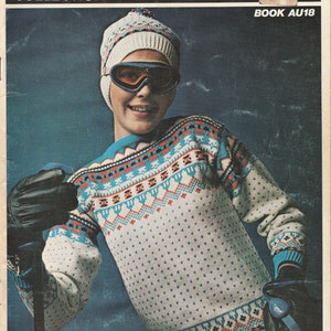 Peut inclure: Un pull à motif blanc et bleu avec un bonnet en tricot blanc et des lunettes de ski. Le pull a un motif géométrique avec des accents rouges et noirs. L'image est tirée d'un livre de modèles de tricot intitulé "Empisal International Knitwear Collection Book Au18".