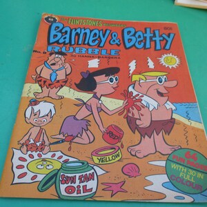 May include: A comic book cover featuring the Flintstones characters Barney and Betty Rubble. The cover shows Barney and Betty on a beach with a sun tan oil bottle and a yellow and red paint can. The text on the cover reads "The Flintstones Neighbors Barney & Betty Rubble by Hanna-Barbera".