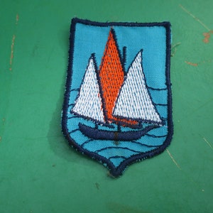 May include: A blue and white embroidered patch featuring a sailboat on a blue background. The sailboat has a white sail and an orange sail.