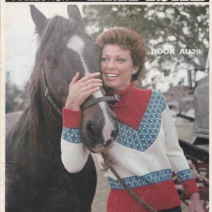 Peut inclure: Une femme portant un pull blanc, bleu et rouge à motifs géométriques tient la tête d'un cheval brun. Le pull a un col en V et des manches longues. La femme sourit et regarde la caméra. L'image est tirée d'un livre de modèles de tricot intitulé "Empisal International Knitwear Collection Book AU29".