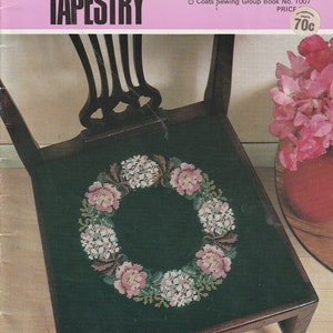 May include: A green chair with a floral tapestry seat featuring a wreath of pink and white flowers. The chair is on a wooden floor and there is a vase of pink flowers in the background.