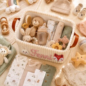 May include: A cream-colored corduroy storage basket with brown leather handles, featuring the embroidered name Fenni, a rabbit face, and a fox face. The basket is filled with baby items including a teddy bear, a bottle, and various textiles.
