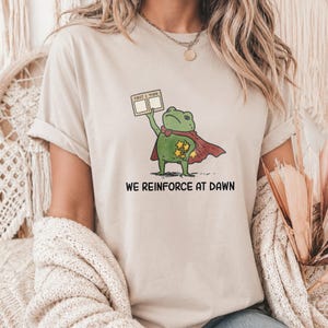 Peut inclure: Un t-shirt beige à manches courtes avec une illustration de grenouille verte portant une cape rouge, tenant un panneau FIRST THEN et des étoiles jaunes. Le texte WE REINFORCE AT DAWN est imprimé en noir sous le dessin de la grenouille.