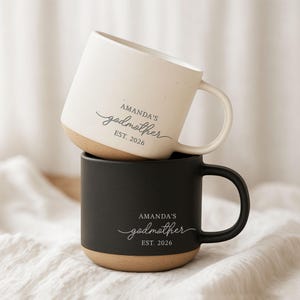 May include: Two ceramic mugs stacked on top of each other. The top mug is off-white with a tan base, and the bottom mug is black with a tan base. Both mugs have the text "AMANDA'S godmother EST. 2026" in a script font.