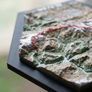 May include: A 3D topographic map with a red line marking a trail. The map features layered contours in shades of brown, green, and white, mounted on a black hexagonal base. The detailed relief shows the terrain's elevation.