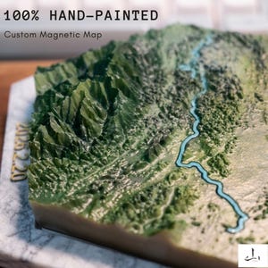 May include: A hand-painted custom magnetic map showing a 3D relief of a mountainous landscape. The map uses green hues for the mountains and a blue river. The text "100% HAND-PAINTED" and "Custom Magnetic Map" are displayed. The map is a unique decorative item.
