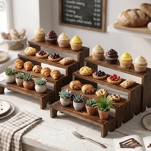 May include: Wooden tiered display stands showcasing an assortment of baked goods, including cupcakes, tarts, and croissants. Small potted succulents add a touch of greenery. The stands are stained a dark brown color.