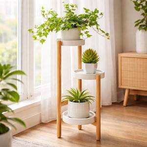 May include: A tiered plant stand featuring three round white shelves supported by light-toned wooden legs. The stand holds a trailing green ivy plant in a white pot on the top shelf, a small succulent in a white pot in the middle, and a spiky plant in a white pot on the bottom shelf.