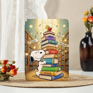 May include: A colorful illustration features Snoopy hugging a stack of books with Woodstock reading on top. The words "I LOVE READING" are on banners. The background shows a library with bookshelves and musical notes.