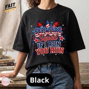 May include: A black t-shirt featuring a graphic with red, white, and blue fireworks and stars. The text reads BOOM Squad IF I RUN YOU RUN in a distressed font with a patriotic flag pattern. The design includes red and blue accents throughout.