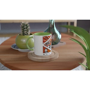 以下が含まれることがあります： 緑色のハンドルと内側を備えた白いセラミックマグ。マグには「Going Hals Mountain」の文字と漫画のクマのグラフィックデザインが施されています。マグは木製のコースターの上にあります。