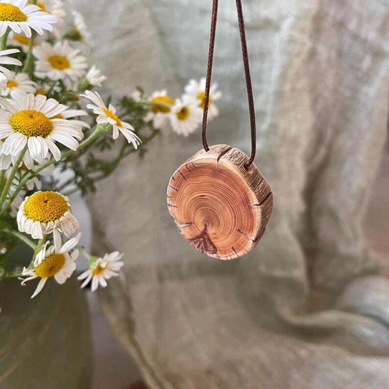 Pode incluir: Um colar com pingente de madeira com um design circular de se&ccedil;&atilde;o transversal de tronco de &aacute;rvore. O pingente &eacute; marrom com an&eacute;is de crescimento vis&iacute;veis, suspenso por um cord&atilde;o marrom. Fundo com margaridas brancas.