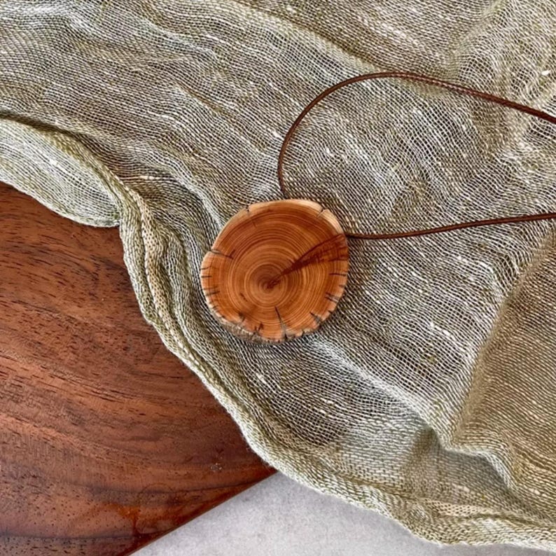 Pode incluir: Um pingente redondo de madeira com um padr&atilde;o de gr&atilde;o natural e um cord&atilde;o de couro marrom. O pingente &eacute; de cor marrom claro com detalhes em marrom escuro, e o cord&atilde;o &eacute; marrom escuro. O pingente &eacute; exibido em um tecido texturizado verde claro.