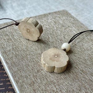 May include: Two wooden flower-shaped pendants with a natural wood grain finish. One pendant is attached to a black cord, and the other is lying on a textured surface. The pendants have a small hole in the centre.