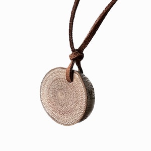 May include: A round pendant necklace with a brown leather cord. The pendant is a light brown color with a textured, patterned surface. The cord is knotted at the top, and the pendant hangs below.