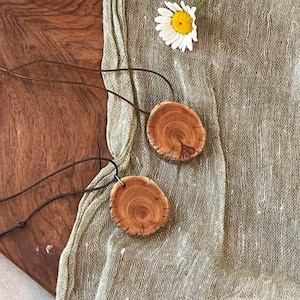Pode incluir: Dois colares com pingentes de madeira com cord&otilde;es de couro castanho. Os pingentes s&atilde;o circulares, cortados de galhos de &aacute;rvores e exibem o gr&atilde;o natural da madeira. Uma margarida branca com centro amarelo repousa sobre um tecido verde.