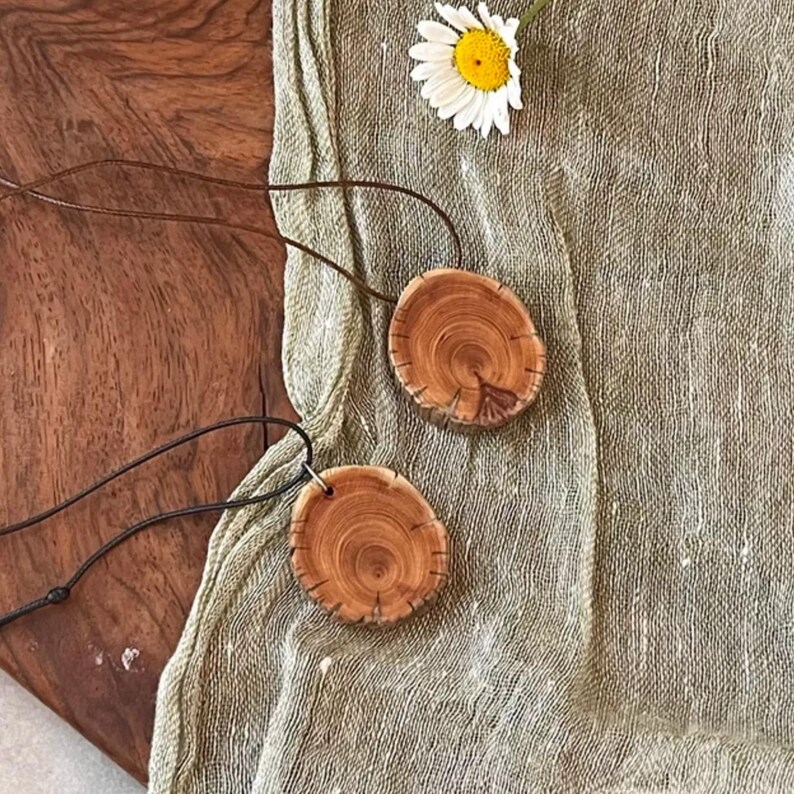 Pode incluir: Dois colares com pingentes de madeira com cord&otilde;es de couro castanho. Os pingentes s&atilde;o circulares, cortados de galhos de &aacute;rvores e exibem o gr&atilde;o natural da madeira. Uma margarida branca com centro amarelo repousa sobre um tecido verde.