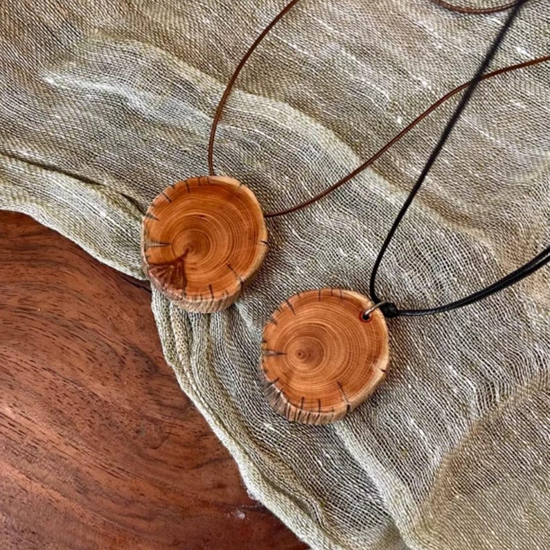 Pode incluir: Dois colares com pingentes de madeira. Cada pingente &eacute; uma fatia circular de madeira, mostrando o gr&atilde;o e a textura naturais. Um colar tem um cord&atilde;o de couro marrom, o outro um cord&atilde;o preto. Os pingentes repousam sobre um tecido bege.