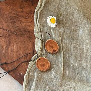Pode incluir: Dois colares com pingentes de madeira com um design de gr&atilde;o de madeira natural. Os pingentes s&atilde;o redondos e presos a cord&otilde;es pretos. Uma margarida branca com centro amarelo repousa sobre um tecido verde claro.