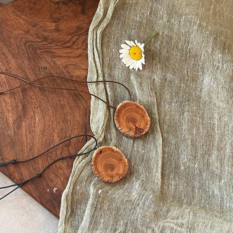 Pode incluir: Dois colares com pingentes de madeira com um design de gr&atilde;o de madeira natural. Os pingentes s&atilde;o redondos e presos a cord&otilde;es pretos. Uma margarida branca com centro amarelo repousa sobre um tecido verde claro.