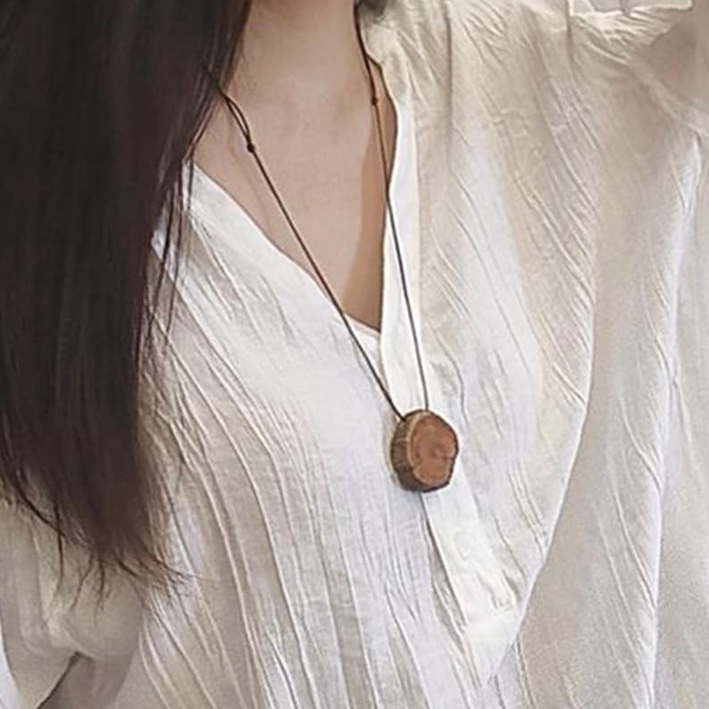 Pode incluir: Um colar com pingente de madeira com um design natural e org&acirc;nico. O pingente &eacute; uma fatia circular de madeira, mostrando o gr&atilde;o e a textura da &aacute;rvore. O cord&atilde;o do colar &eacute; marrom escuro, complementando os tons quentes do pingente. O colar &eacute; usado sobre uma blusa branca.
