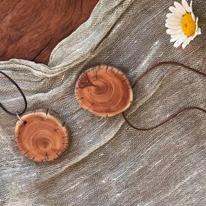 Pode incluir: Dois colares com pingentes de madeira com uma est&eacute;tica natural e r&uacute;stica. Cada pingente &eacute; uma fatia circular de madeira, mostrando os an&eacute;is de crescimento da &aacute;rvore. Os pingentes s&atilde;o conectados a um cord&atilde;o de couro marrom escuro. Uma flor de margarida ao fundo.