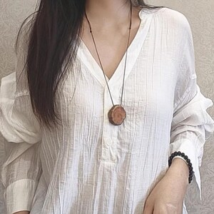 Pode incluir: Um colar com pingente de madeira com um design circular de se&ccedil;&atilde;o transversal. O pingente &eacute; de cor marrom quente e pende de um cord&atilde;o preto. O colar &eacute; usado sobre uma camisa branca de manga comprida.