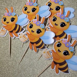 K&ouml;nnte beinhalten: Sechs Cupcake-Topper mit Cartoon-Bienen. Jede Biene hat orange und schwarze Streifen, blaue Fl&uuml;gel und einen Blumenkranz. Sie halten kleine beige Gie&szlig;kannen und sind an Holzst&auml;bchen befestigt.