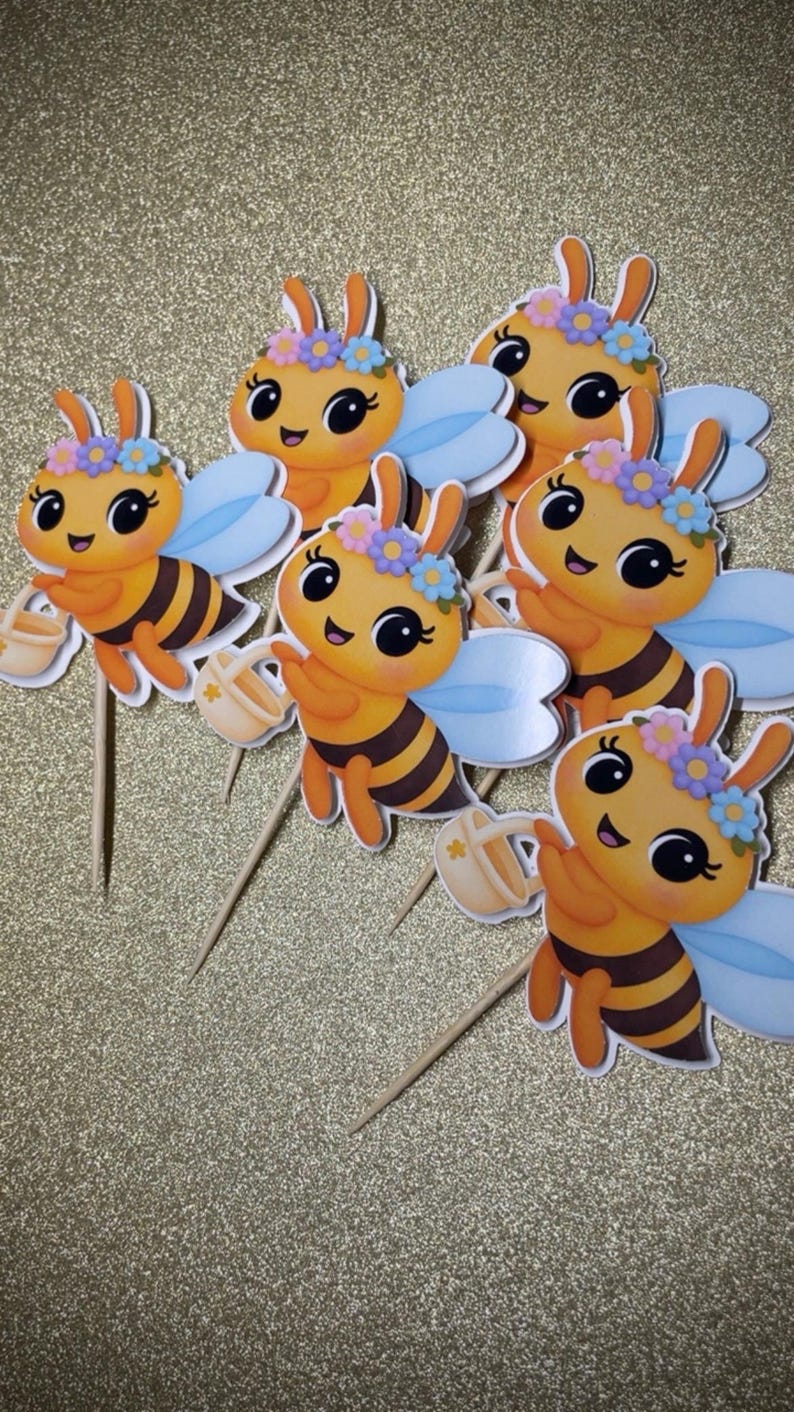 K&ouml;nnte beinhalten: Sechs Cupcake-Topper mit Cartoon-Bienen. Jede Biene hat orange und schwarze Streifen, blaue Fl&uuml;gel und einen Blumenkranz. Sie halten kleine beige Gie&szlig;kannen und sind an Holzst&auml;bchen befestigt.