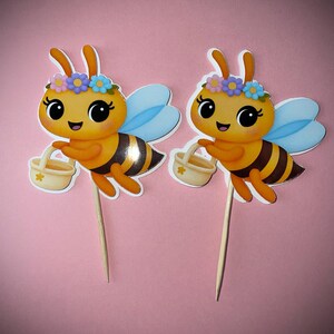 K&ouml;nnte beinhalten: Zwei Bienen-Kuchenaufs&auml;tze. Jede Biene hat einen gelben K&ouml;rper mit schwarzen Streifen, blaue Fl&uuml;gel und einen Blumenkranz. Sie halten kleine K&ouml;rbe und sind an Holzspie&szlig;en befestigt. Der Hintergrund ist einfarbig rosa.