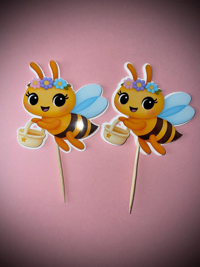 K&ouml;nnte beinhalten: Zwei Bienen-Kuchenaufs&auml;tze. Jede Biene hat einen gelben K&ouml;rper mit schwarzen Streifen, blaue Fl&uuml;gel und einen Blumenkranz. Sie halten kleine K&ouml;rbe und sind an Holzspie&szlig;en befestigt. Der Hintergrund ist einfarbig rosa.