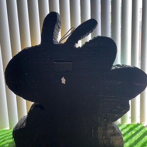 May include: A large, black rabbit-shaped pi&ntilde;ata, crafted from layered paper. It features long ears and a small white flower. The pi&ntilde;ata rests on a patch of green artificial grass.